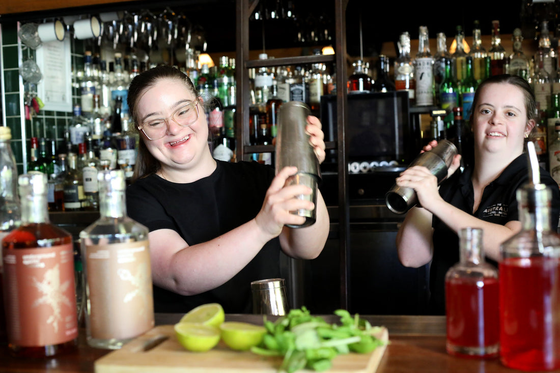 Two young women with Down syndrome smile and shake cocktail shakers behind a bar, surrounded by fresh ingredients and Underground Spirits bottles. Their joyful expressions highlight inclusion and empowerment in a welcoming social setting.