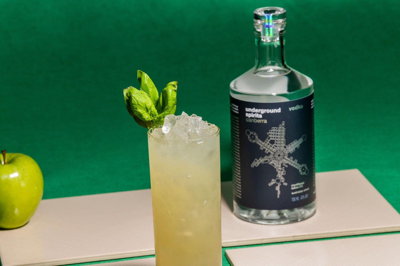 A bottle of Underground Spirits Canberra Vodka, accompanied by a refreshing cocktail garnished with basil leaves and ice, sits elegantly next to a green apple against a vibrant green background, emphasising the natural, high-quality ingredients used by Underground Spirits